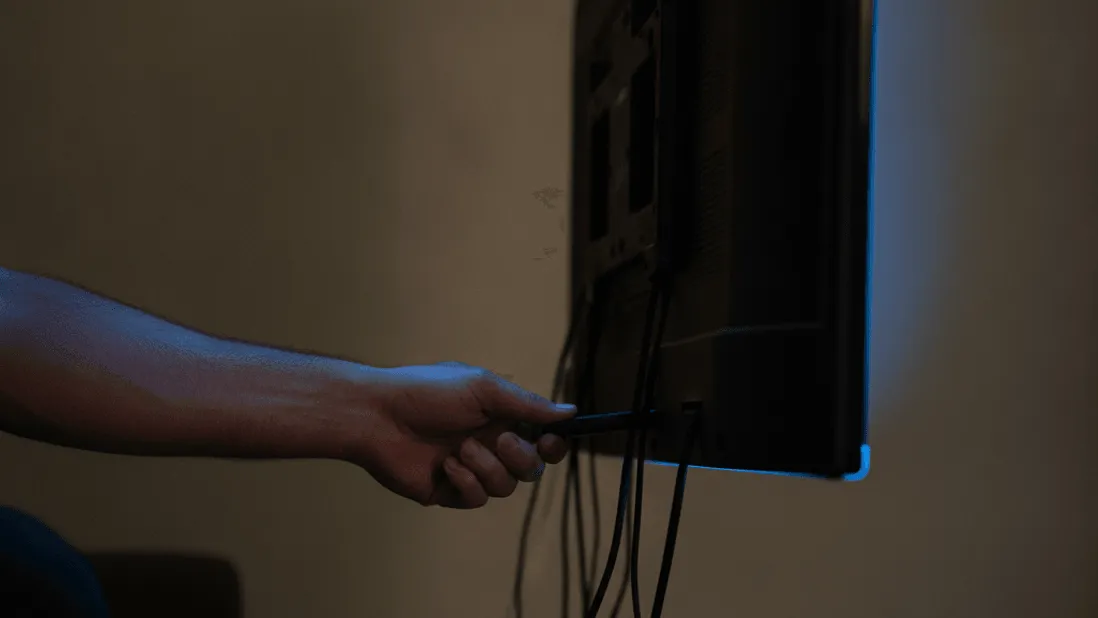 A hand plugs a streaming stick into the back of a wall-mounted television in a dimly lit room.