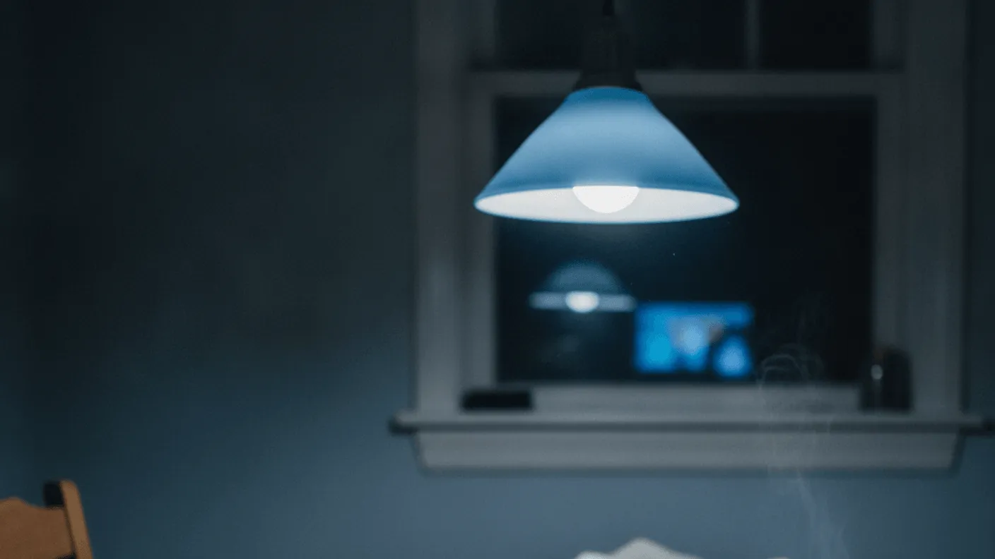 A kitchen table at night with a receipt, coins, calculator, and a steaming mug under a blue-cast lamp.