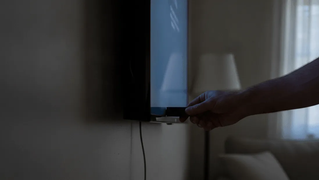 A hand inserts a streaming stick into a wall-mounted television in a dimly lit room with a window and a sofa.