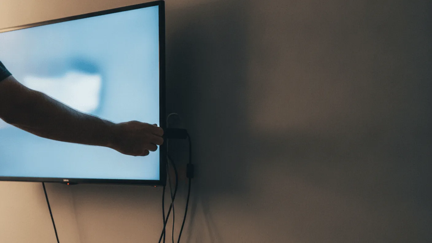 A hand plugs a small device into the back of a wall-mounted television in a dimly lit room.