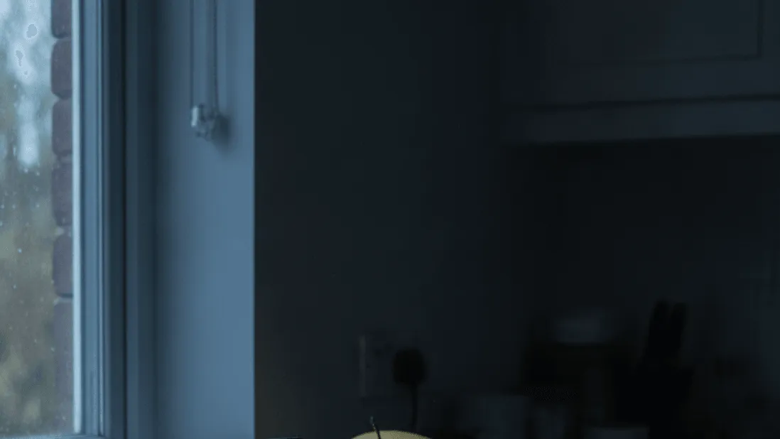 A tablet propped against a fruit bowl on a kitchen counter in low light, with a hand holding a mug of tea.