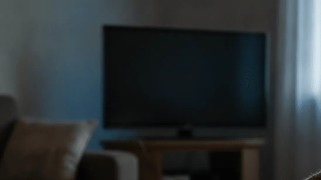 A hand reaches for a remote control on a coffee table beside a streaming box in a dimly lit room.