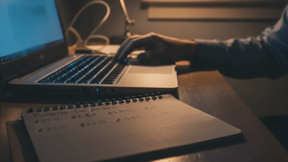 A man works on a laptop at a desk, with a notepad of numbers beside him, illuminated by a desk lamp and fading window light.