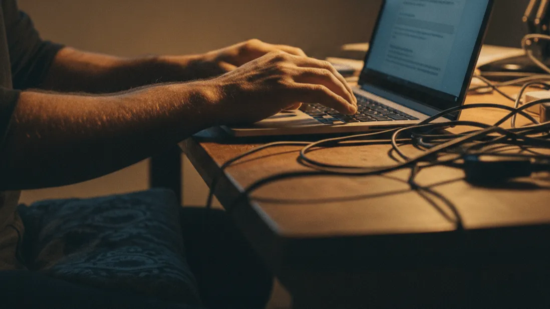 A person types on a laptop keyboard at a desk, illuminated by a desk lamp and screen light, with cables scattered.