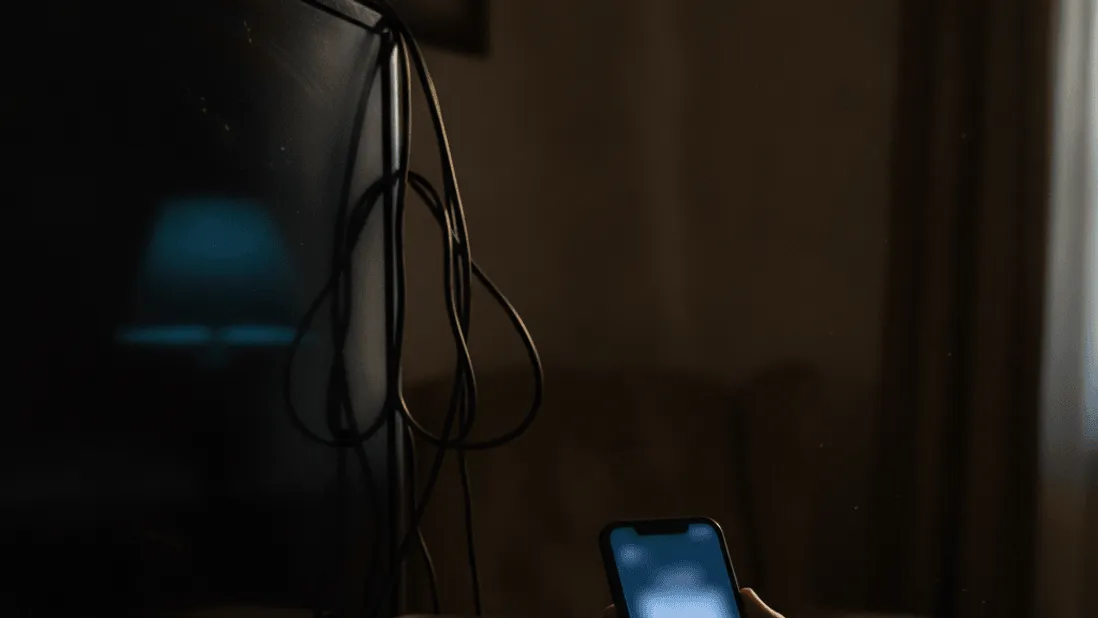 A hand holds a smartphone near a partially connected streaming device in dim, lived-in room.