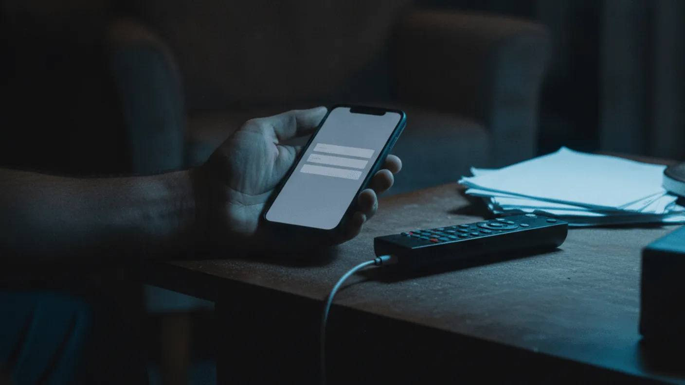 A hand holds a smartphone with a blank login screen on a wooden table beside a remote control under lamp light.